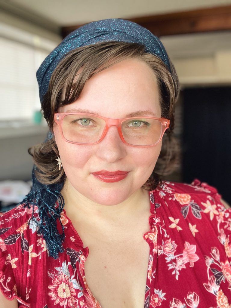 Photo of woman in mid-30s with medium brown hair, coral glasses, dangling star earrings, red dress, and peacock blue scarf in the hair.